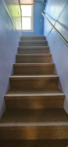 Carpeted staircase with an integrated handrail, illuminated by an overhead window with a blind