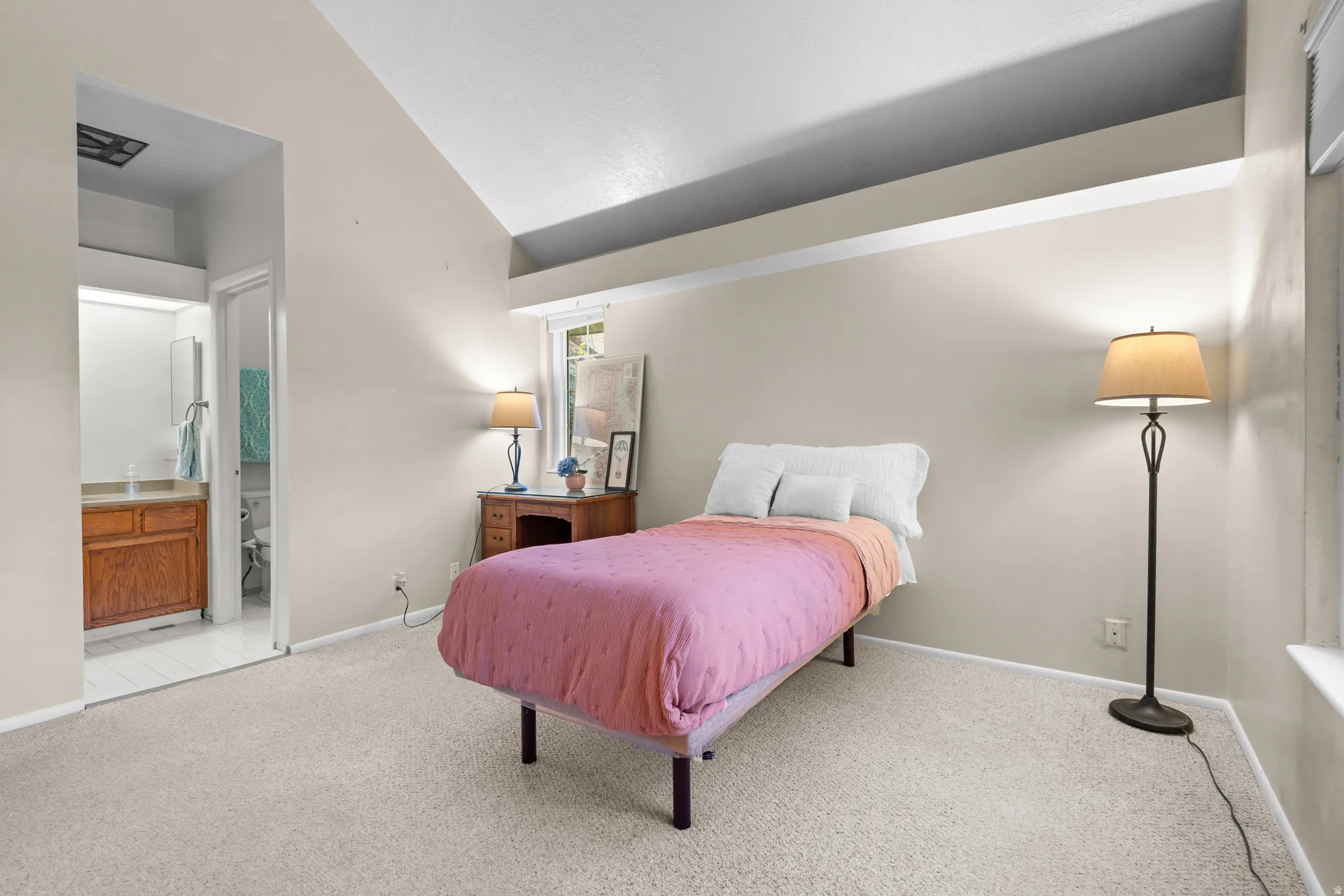 Spacious carpeted room featuring vaulted ceilings, a window providing natural light, and an adjoining bathroom with a wood-finish vanity