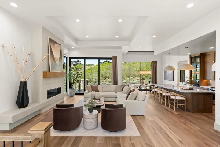 Open-concept living space featuring wood-finish flooring, a linear fireplace with a stone surround and wood mantel, large glass sliding doors, recessed lighting, and a kitchen island with seating