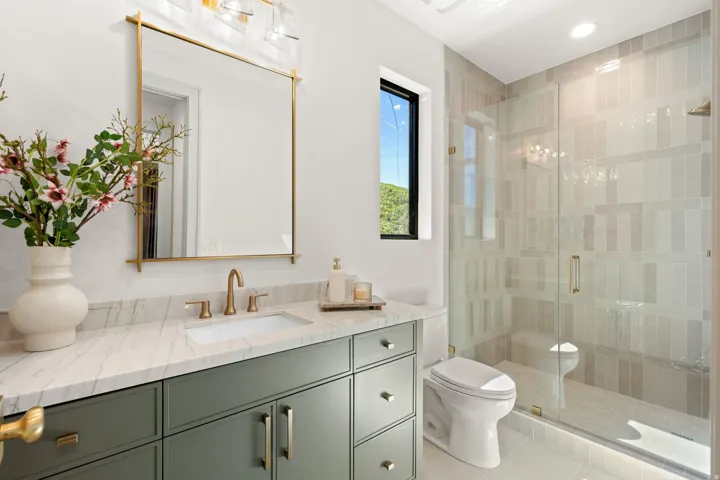 Bathroom featuring a sage green vanity with a white countertop and gold-toned hardware, a frameless glass shower enclosure with vertical tile accents, a rectangular window with black trim, a gold-framed mirror, and a contemporary vanity light fixture