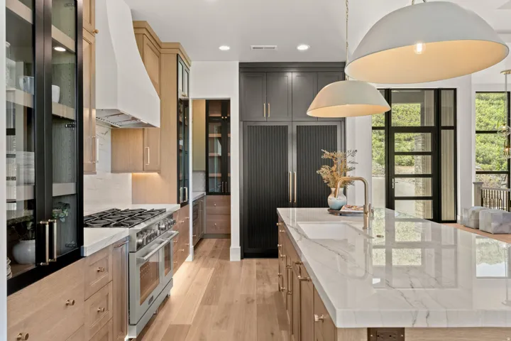 Gourmet kitchen featuring a large central island with an undermount sink and brass faucet, light wood-finish cabinetry with brass hardware, integrated stainless steel appliances, a white range hood, and black-framed glass paneled doors