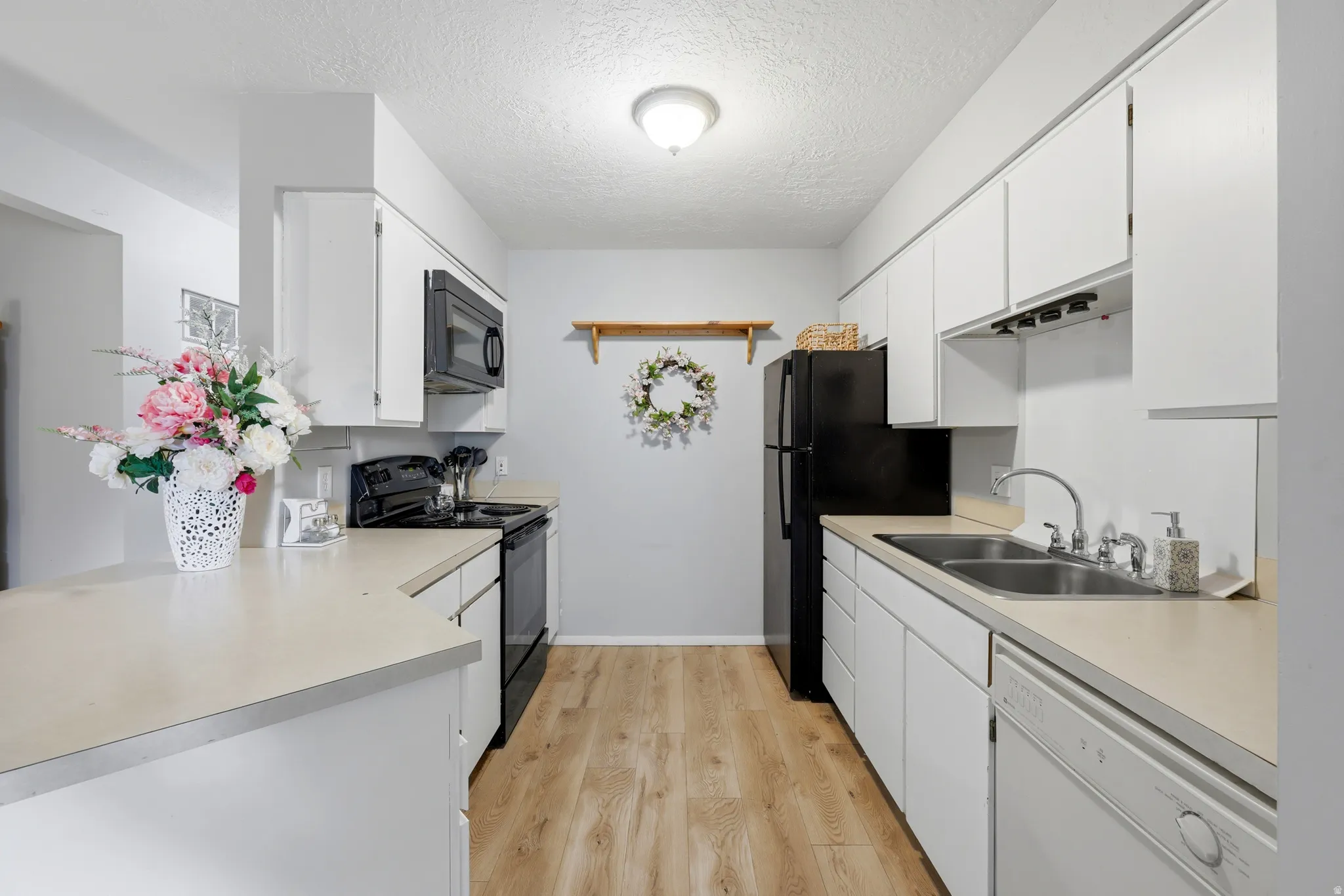Bright kitchen featuring white cabinetry, light-toned countertops, wood-finish flooring, a double basin stainless steel sink, and a full suite of black appliances