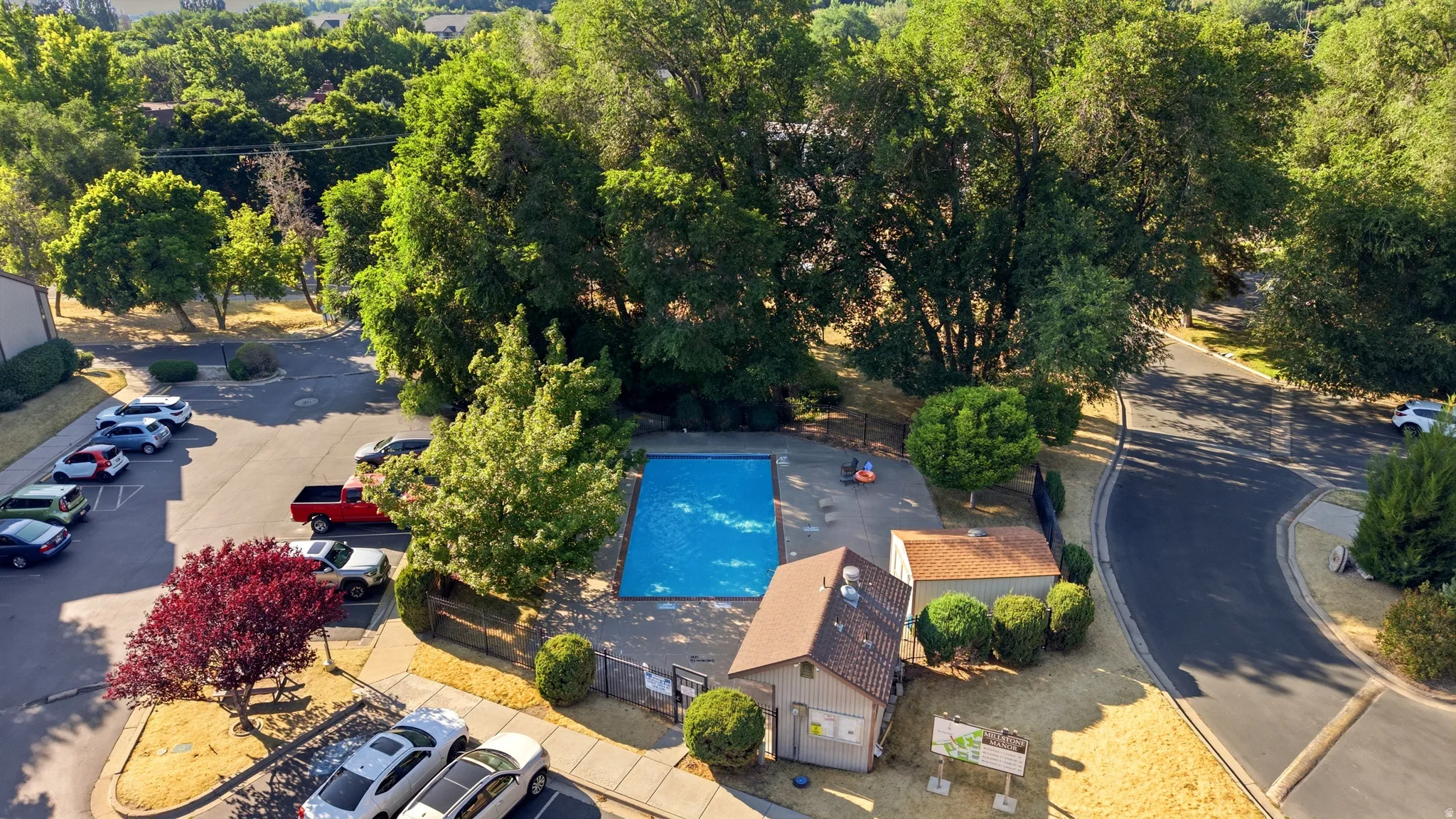 Outdoor swimming pool surrounded by mature trees, featuring a nearby building with a shingled roof, paved parking areas, and surrounding green landscaping