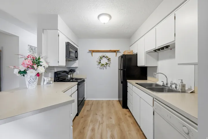 Bright kitchen featuring white cabinetry, light-toned countertops, wood-finish flooring, a double basin stainless steel sink, and a full suite of black appliances