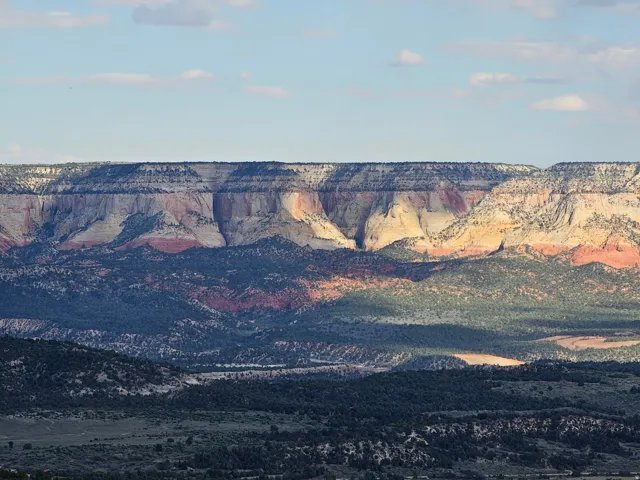 Expansive natural landscape featuring stratified rock formations with varied coloration, brush-covered hills, and a broad sky