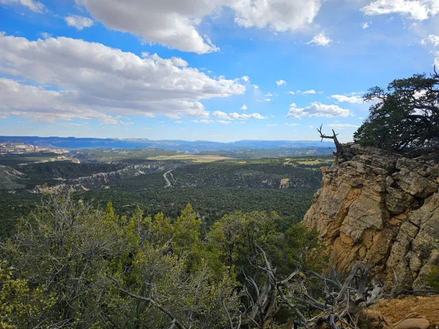 Expansive natural terrain featuring a prominent rock cliff, distant green valleys, and a winding road through the landscape