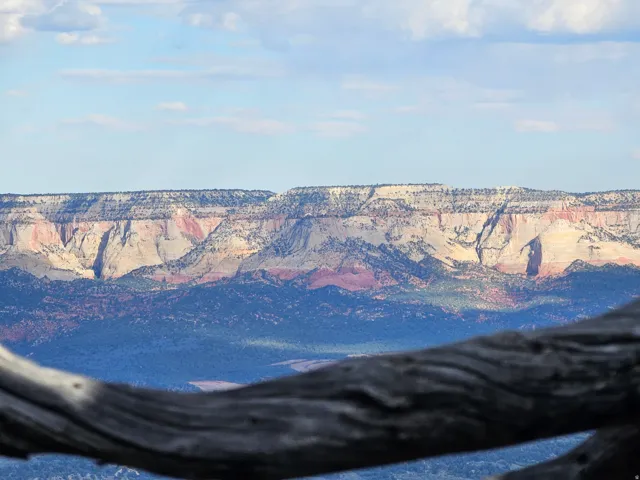Expansive natural landscape featuring distant layered mountains with varied rock formations and colors, set against a light blue sky with scattered clouds