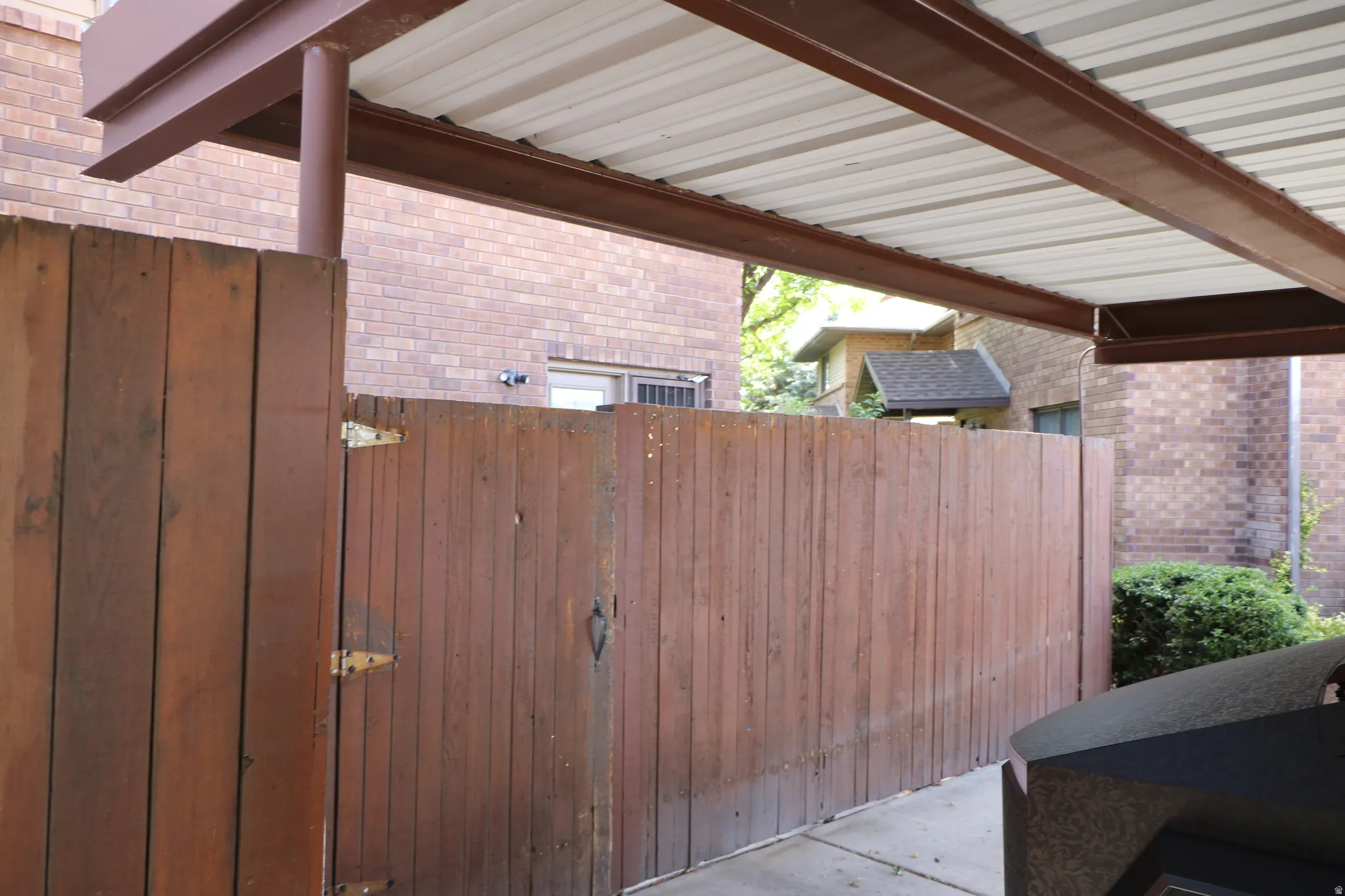 Covered carport with a corrugated metal roof and exposed brown metal beams, featuring a wood-panel fence with visible hinges and a brick exterior building in the background