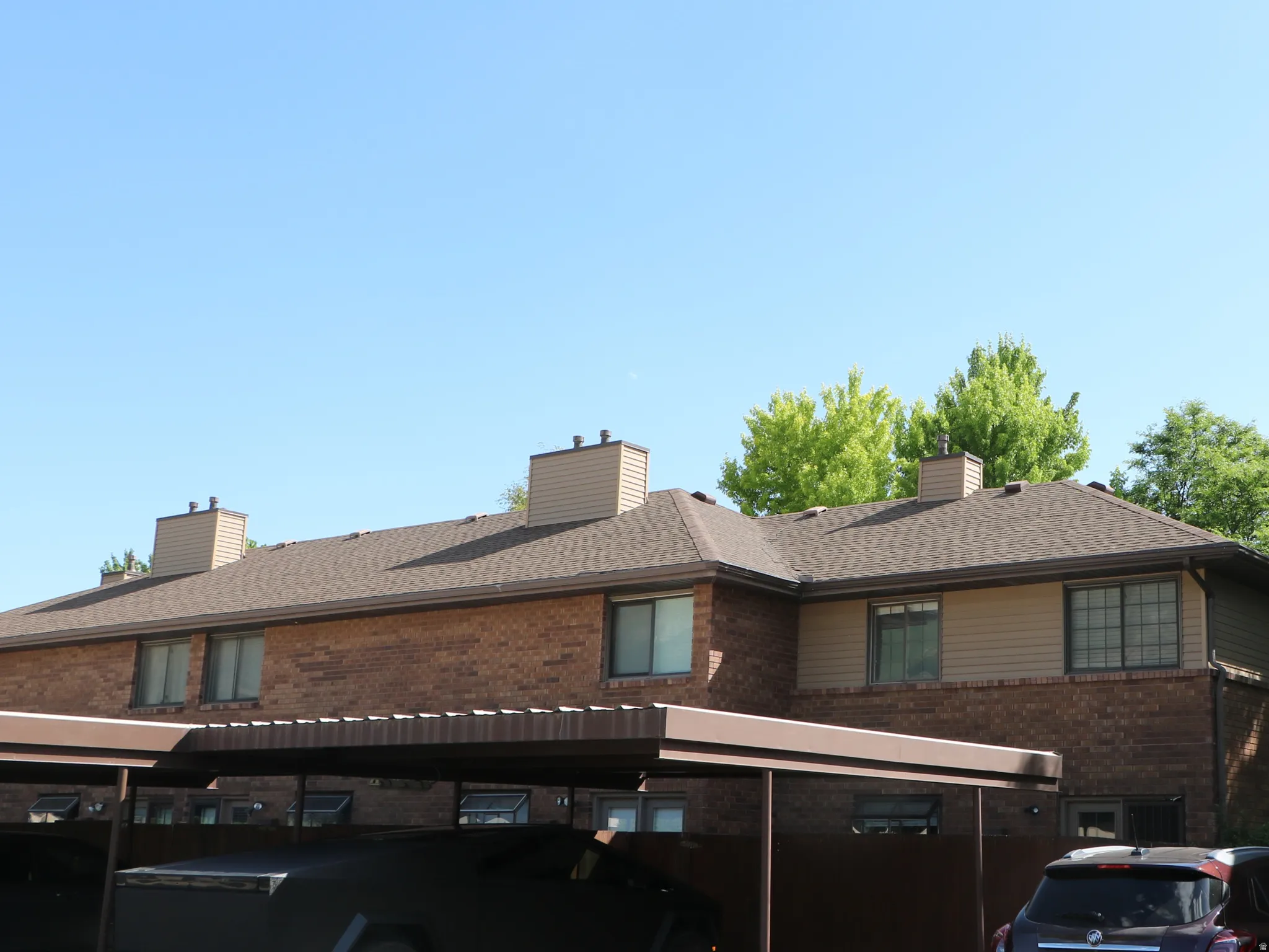 Brick townhouse exterior with multiple windows, individual covered parking spaces, a shingled roof, and a brick chimney