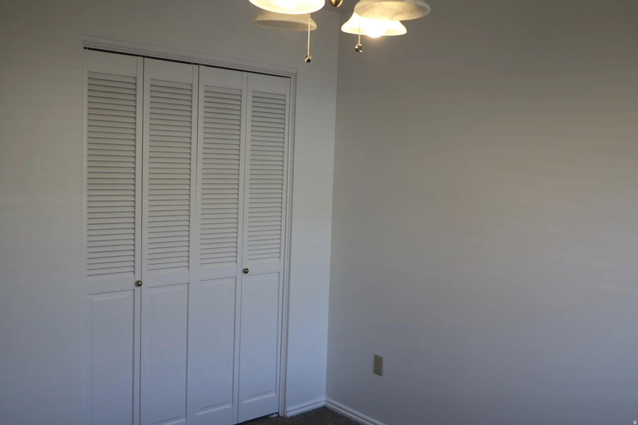 Wall-to-wall louvered bi-fold closet doors with brass-finish hardware, light neutral wall paint, and a ceiling-mounted light fixture with frosted glass shades