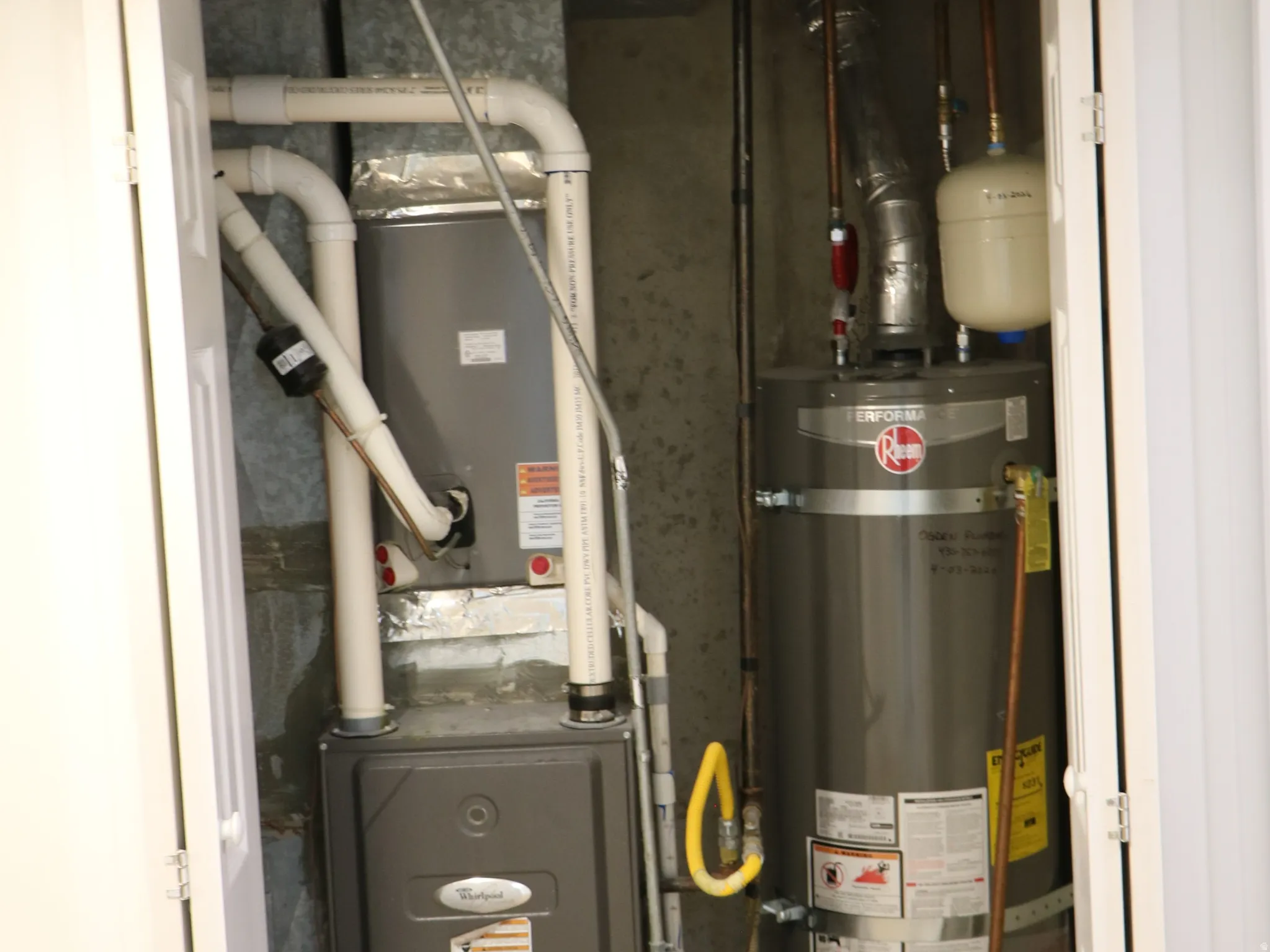 Utility closet housing a water heater and HVAC unit, featuring white louvered bifold doors and textured tile flooring