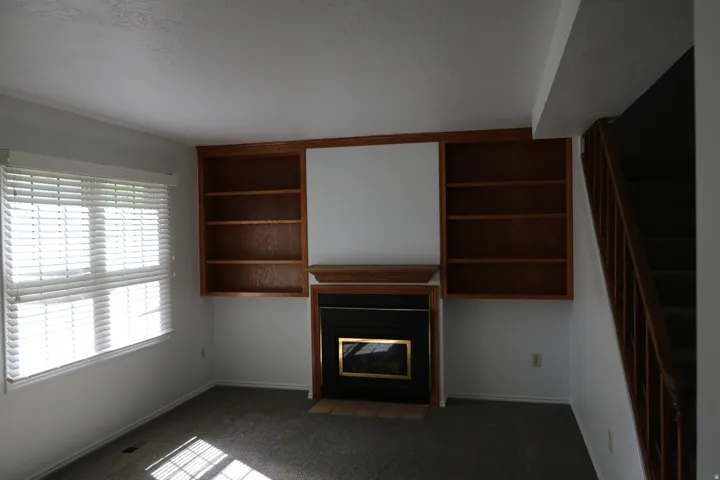 Living area featuring a fireplace with a brass-trimmed insert, flanked by built-in wooden shelving units, and a window with white blinds