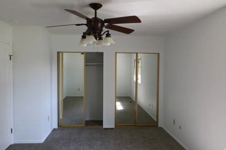 Bedroom featuring a ceiling fan with integrated lighting, carpet flooring, a white paneled door, and two mirrored sliding closet doors with gold-finish frames