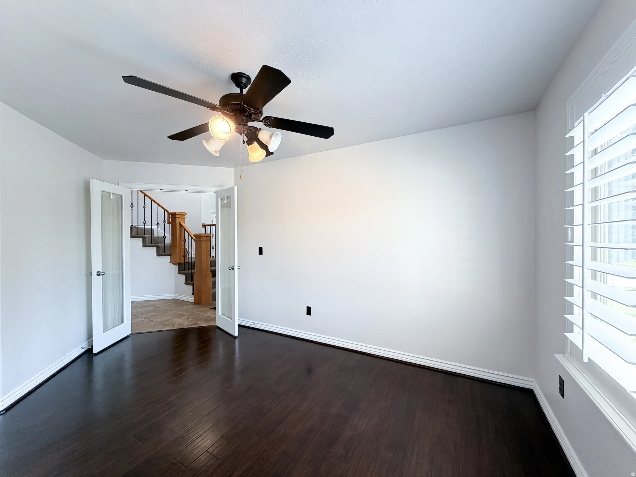 Dark wood-finish flooring with white baseboards, a ceiling fan with integrated lighting, and white plantation shutters on the window