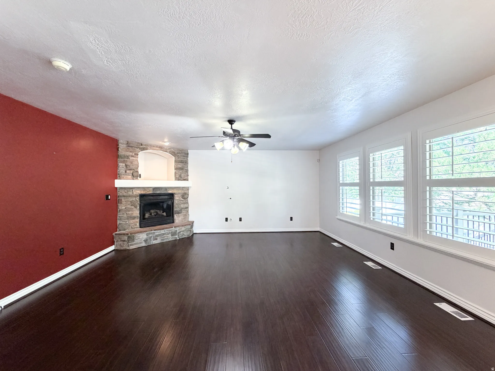 Stone-faced fireplace with hearth, dark wood-finish flooring, ceiling fan with integrated lighting, and multiple windows with plantation shutters