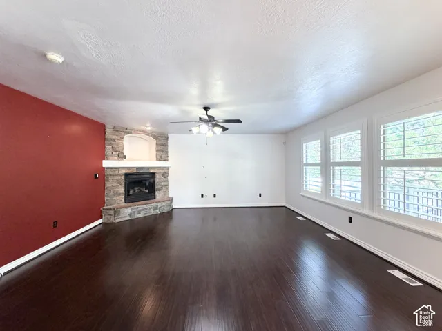 Stone-faced fireplace with hearth, dark wood-finish flooring, ceiling fan with integrated lighting, and multiple windows with plantation shutters