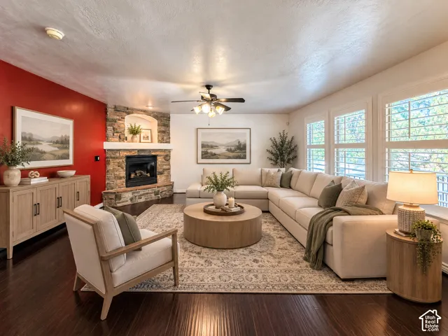 Stone-clad fireplace with hearth, dark wood-finish flooring, ceiling fan, and multiple windows with plantation shutters