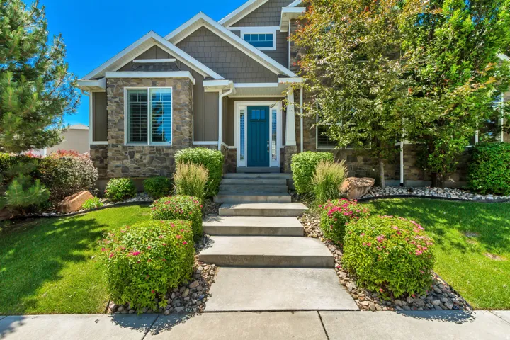 Craftsman-style home featuring stone and siding exterior, a teal front door, concrete walkway with steps, manicured shrubbery, and mature trees