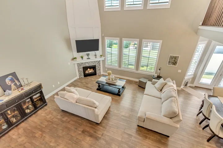 Vaulted living room with wood-finish flooring, a stone-clad fireplace, and multiple windows featuring plantation shutters