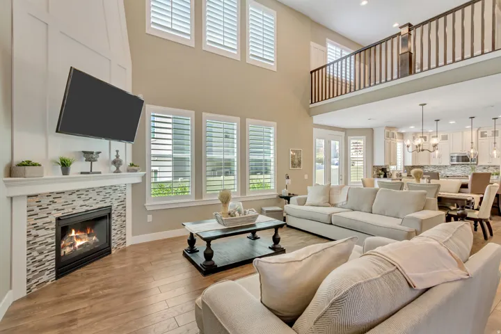 Two-story living area with wood-finish flooring, a contemporary fireplace featuring a mosaic tile surround and white mantel, and multiple windows with plantation shutters