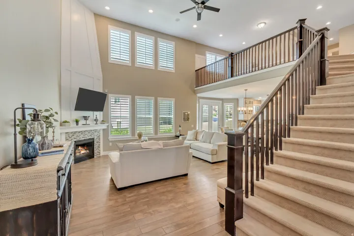 Expansive living room featuring wood-finish flooring, a two-story fireplace with board and batten paneling, multiple windows with plantation shutters, a carpeted staircase with dark wood banisters, and an upper-level balcony overlooking the space