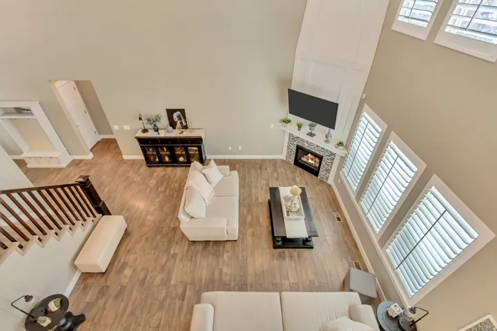 Open-concept living space featuring wood-finish flooring, a stone-faced fireplace with a white mantel, and tall windows with plantation shutters
