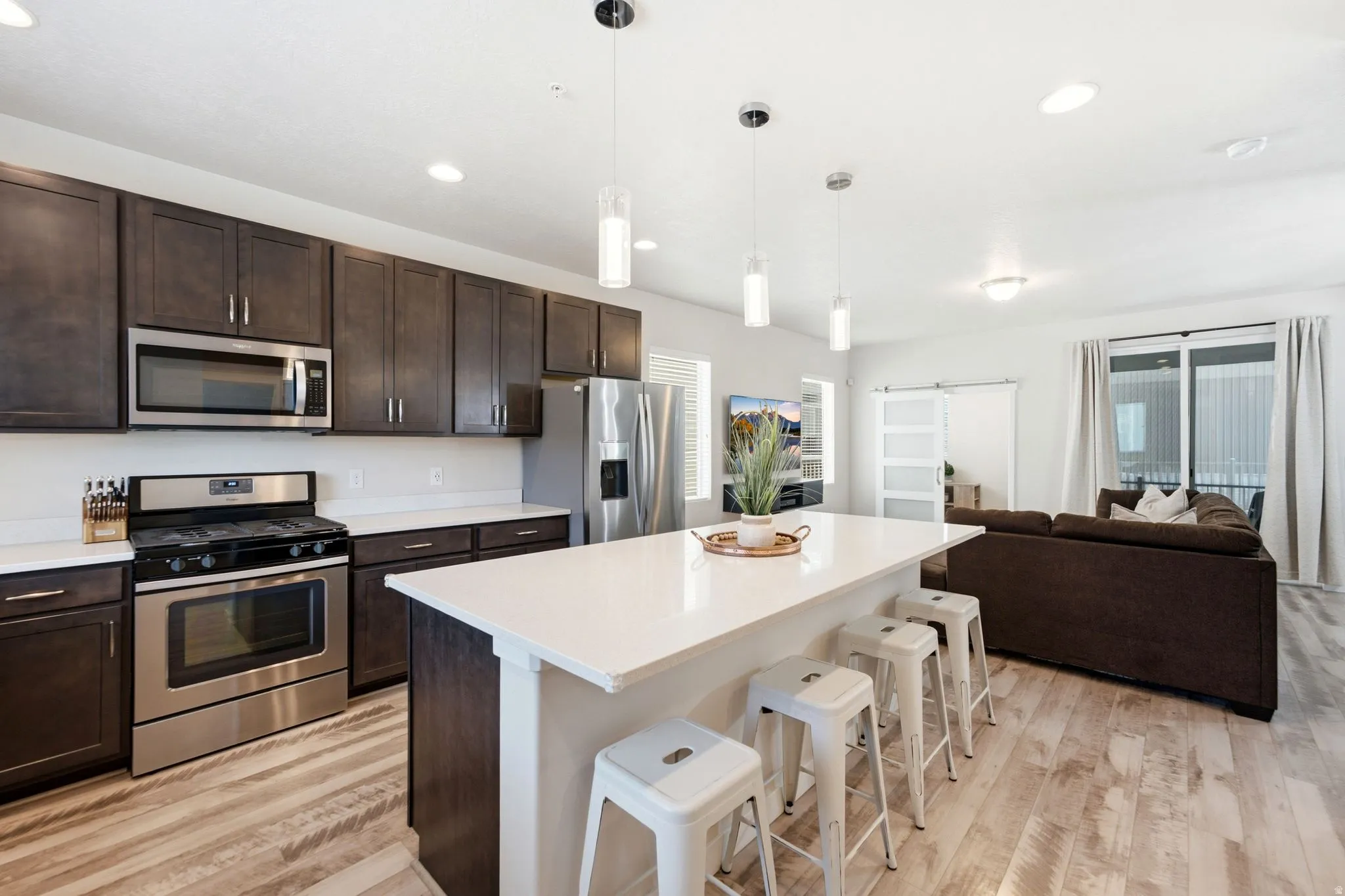 Open-concept kitchen featuring dark wood cabinetry, stainless steel appliances, a white countertop island with pendant lighting, and wood-finish flooring