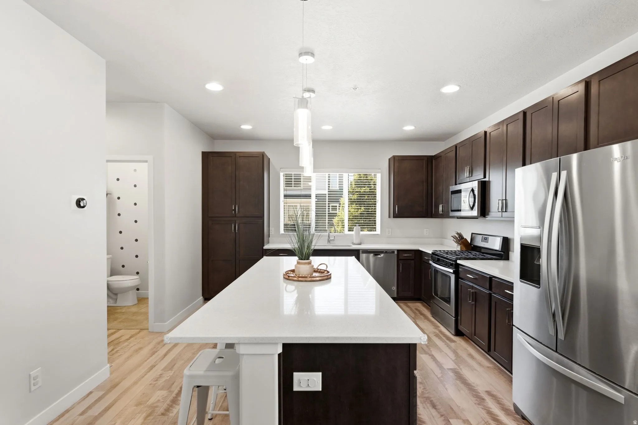 Modern kitchen featuring an expansive center island with a white countertop, dark wood cabinetry, stainless steel appliances, recessed lighting, and wood-finish flooring