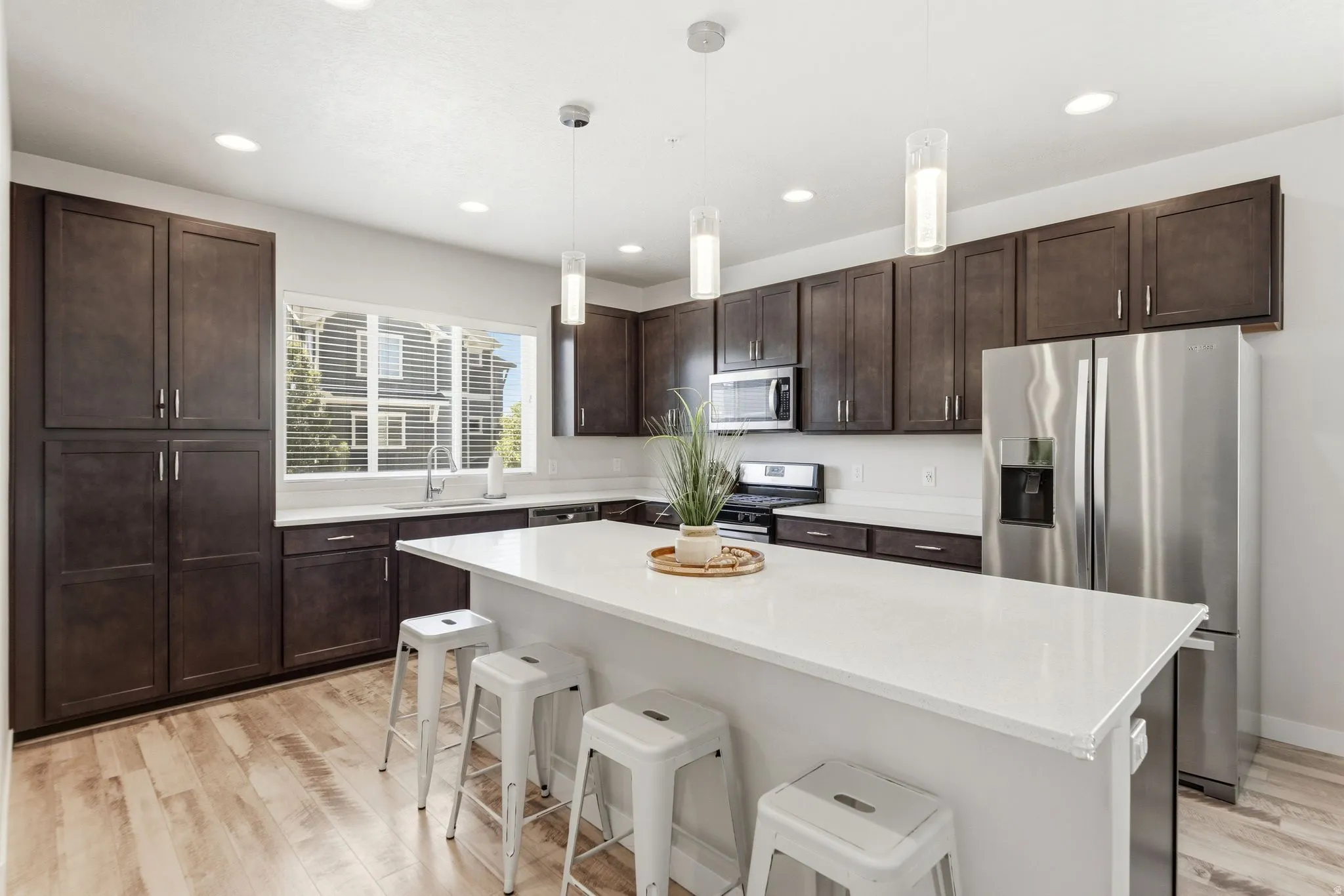 Spacious kitchen featuring an extended center island with seating, dark wood cabinetry, stainless steel appliances, light-toned wood-finish flooring, and ample recessed lighting