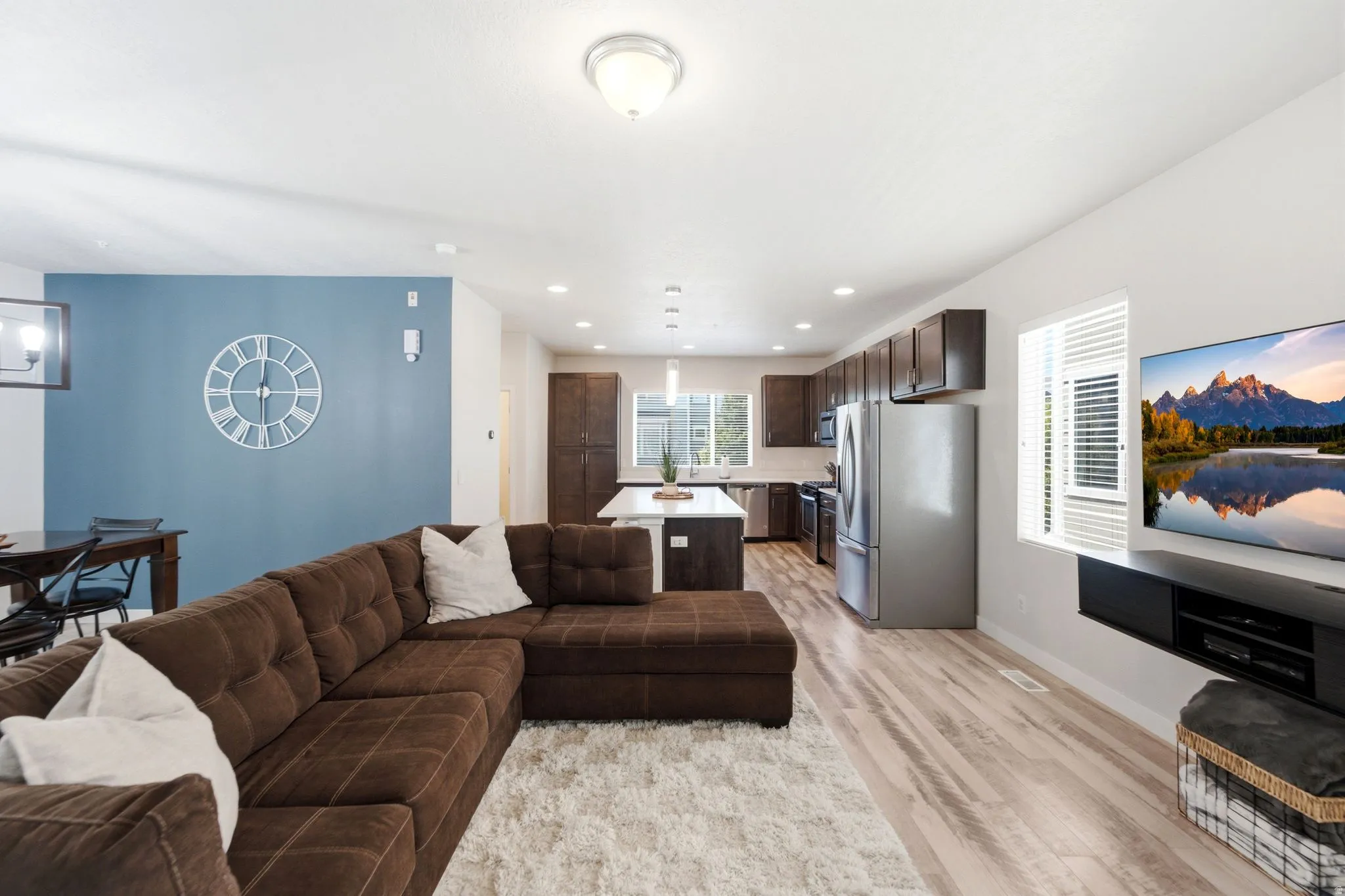 Open-concept living area featuring light wood-finish flooring, recessed lighting, a kitchen with dark cabinetry, stainless steel appliances, and a white island with seating