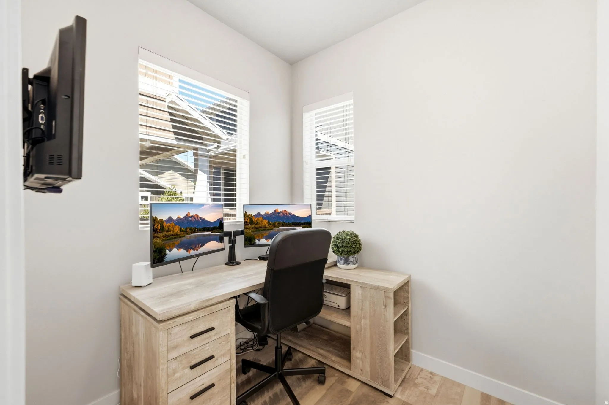 Corner room with two windows featuring white blinds, light wood-finish flooring, and a wall-mounted bracket