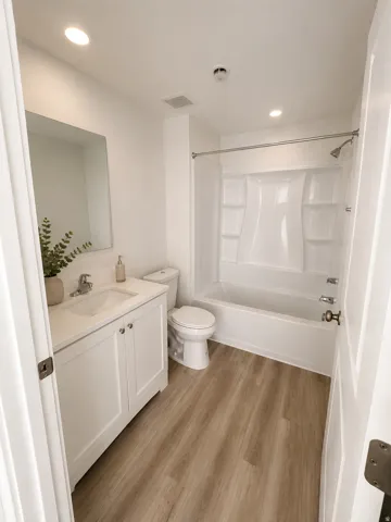 Contemporary bathroom featuring a white vanity with integrated sink, a rectangular mirror, a toilet, a tub with a shower kit, and wood-finish flooring