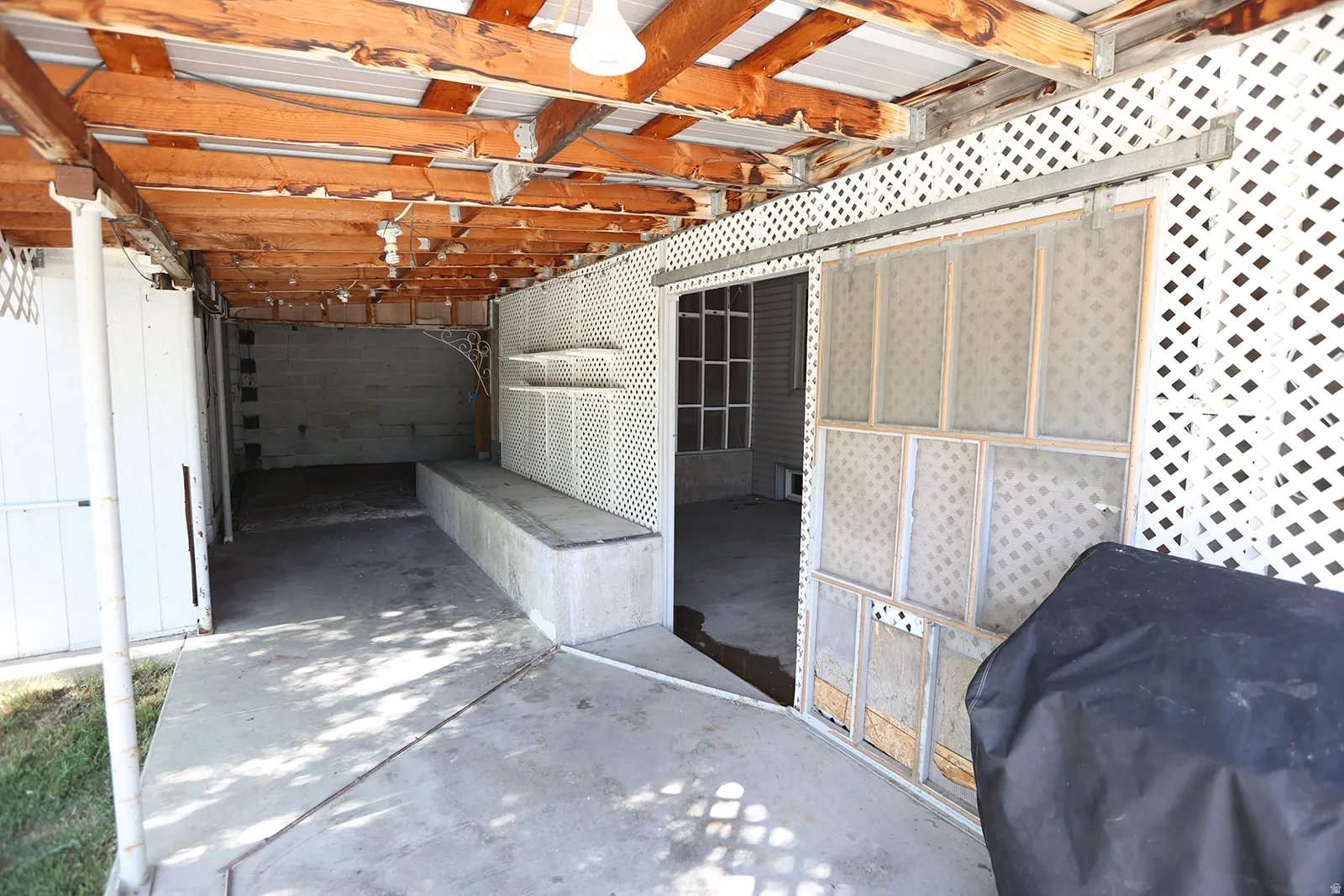 Covered outdoor area featuring a concrete floor, exposed wood beam ceiling, white lattice perimeter, and a built-in concrete bench