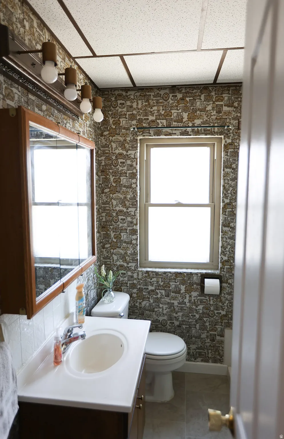 Bathroom featuring a single window, a drop ceiling, patterned wallpaper, a vanity with an integrated sink, and a mirrored medicine cabinet
