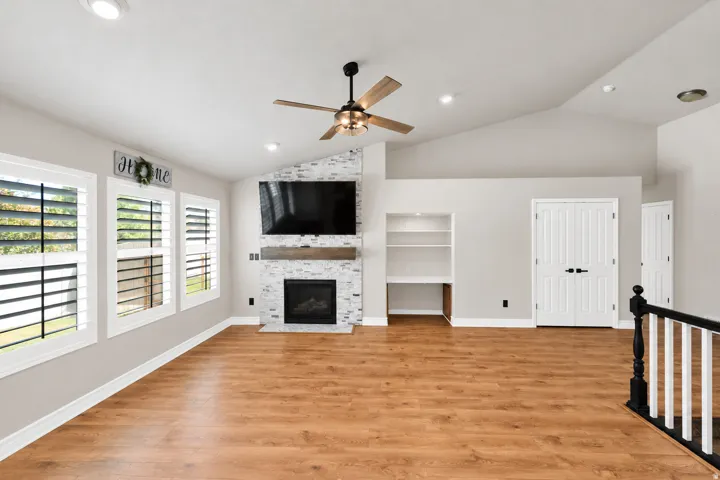 Vaulted ceiling with recessed lighting, wood-finish flooring, fireplace with stone surround, custom built-in shelving, and white plantation shutters