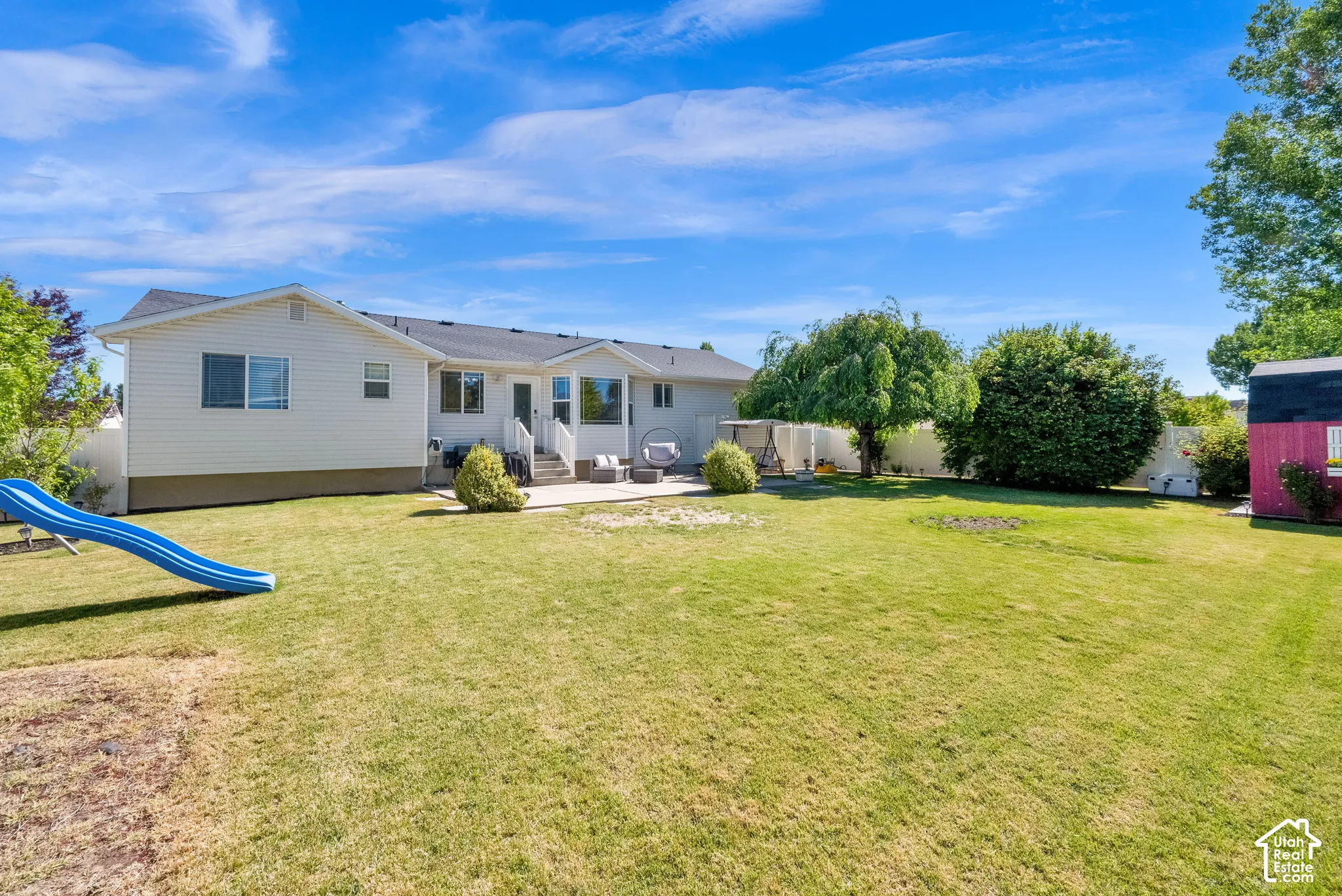 Expansive grassy backyard featuring a concrete patio, mature trees, white vinyl fencing, and a detached storage shed