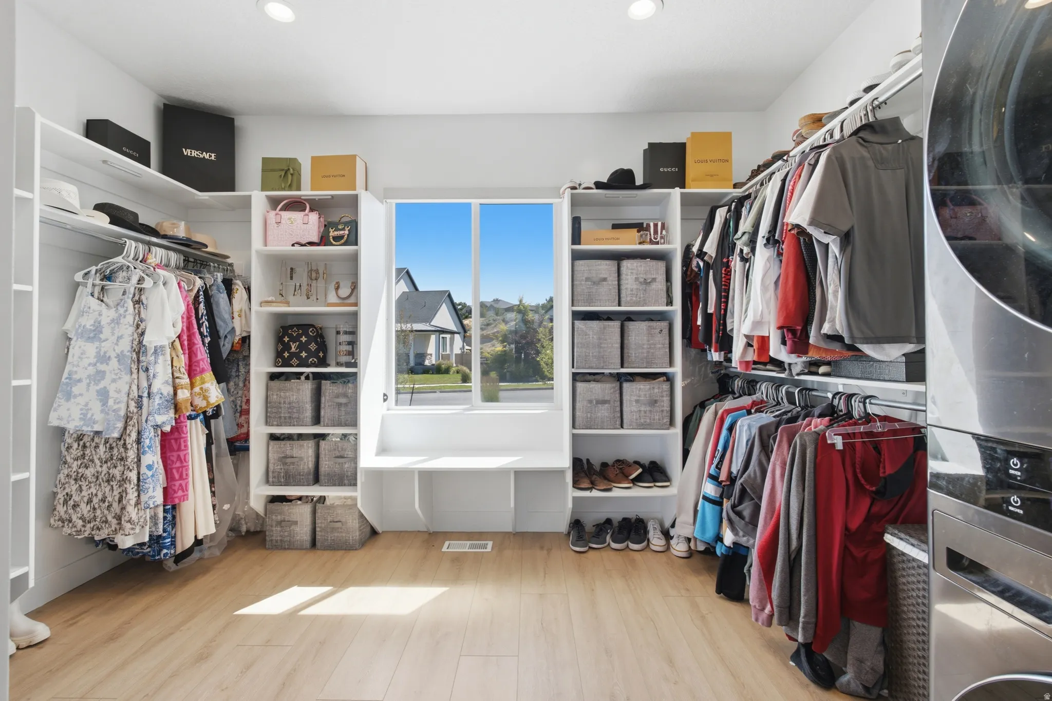 Expansive walk-in closet featuring extensive built-in shelving, hanging rods, light wood-finish flooring, a window with a built-in bench, and recessed lighting