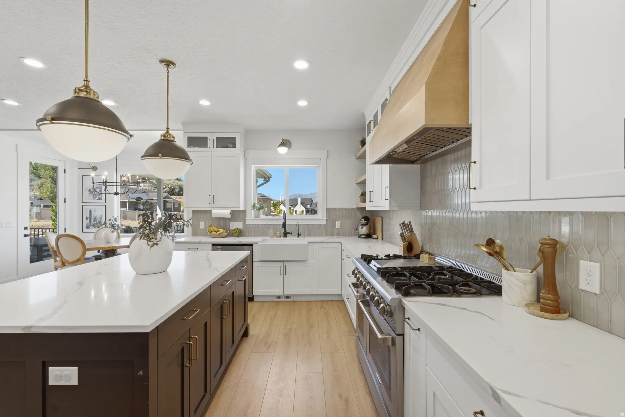 Spacious kitchen featuring an expansive island with a white countertop, two pendant lights, wood-finish flooring, a farmhouse sink, and a range with a custom wood hood