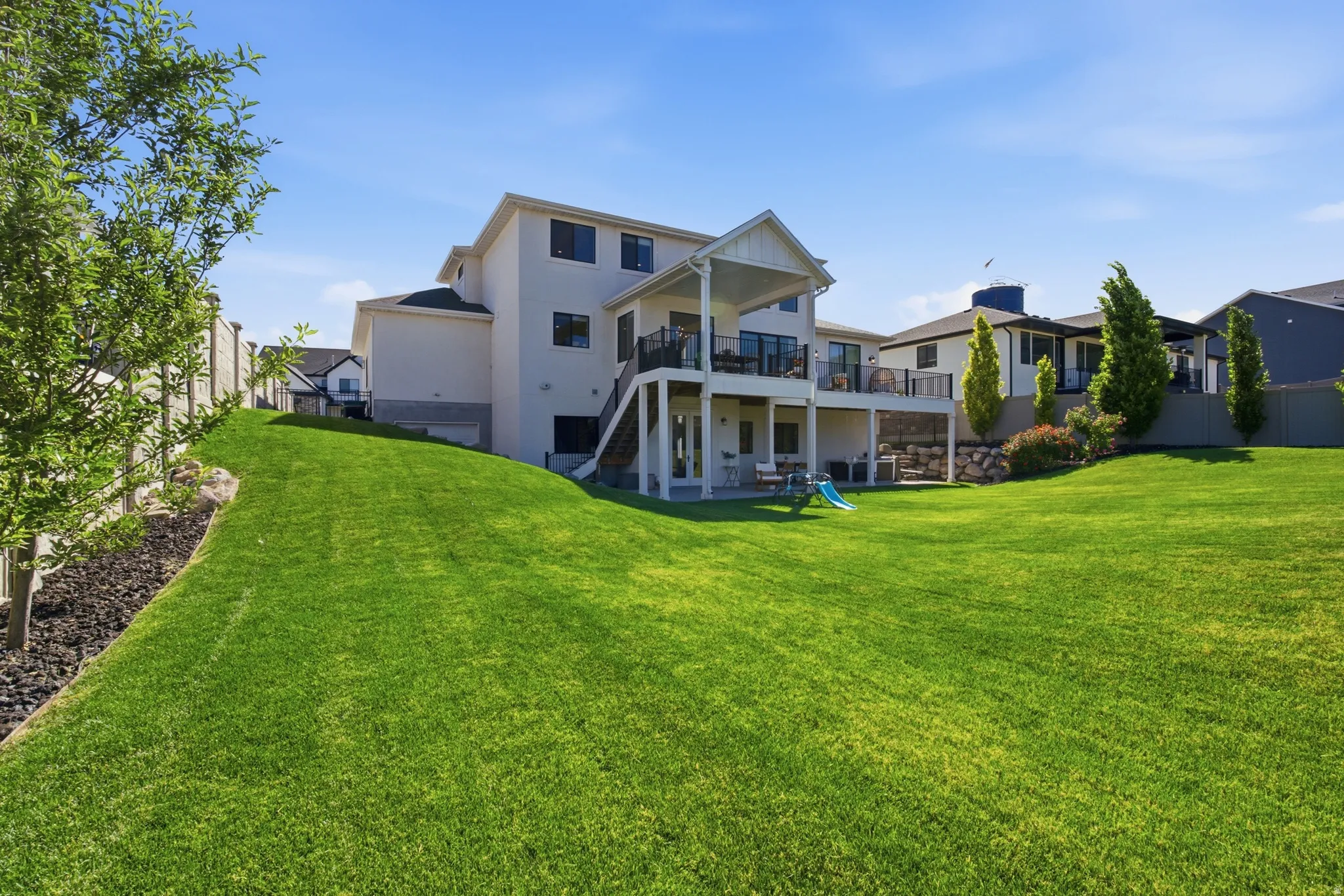 Spacious lawn backing to a multi-story residence with a covered balcony, lower-level patio, and retaining wall landscaping