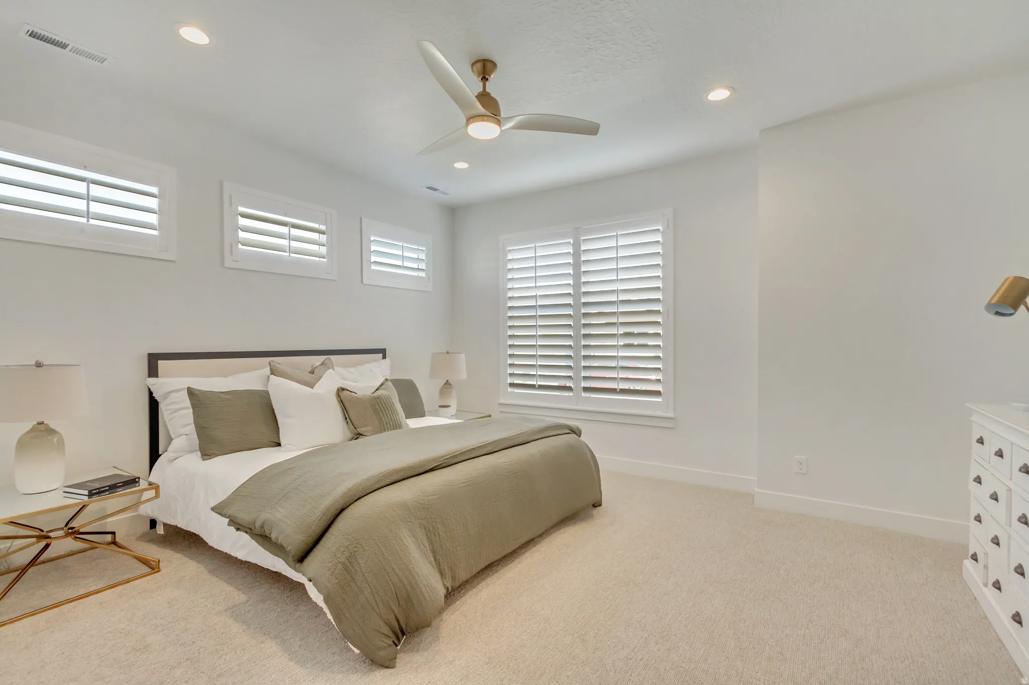 Carpeted room featuring a white upholstered headboard, multiple windows with white plantation shutters, a ceiling fan with integrated lighting, and recessed ceiling lights
