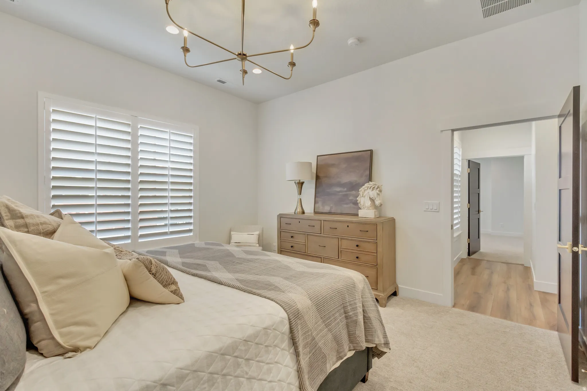 Bedroom featuring white plantation shutters, a six-light brass chandelier, light-toned carpet flooring, a wood-finish dresser with multiple drawers, and a dark wood-finish interior door