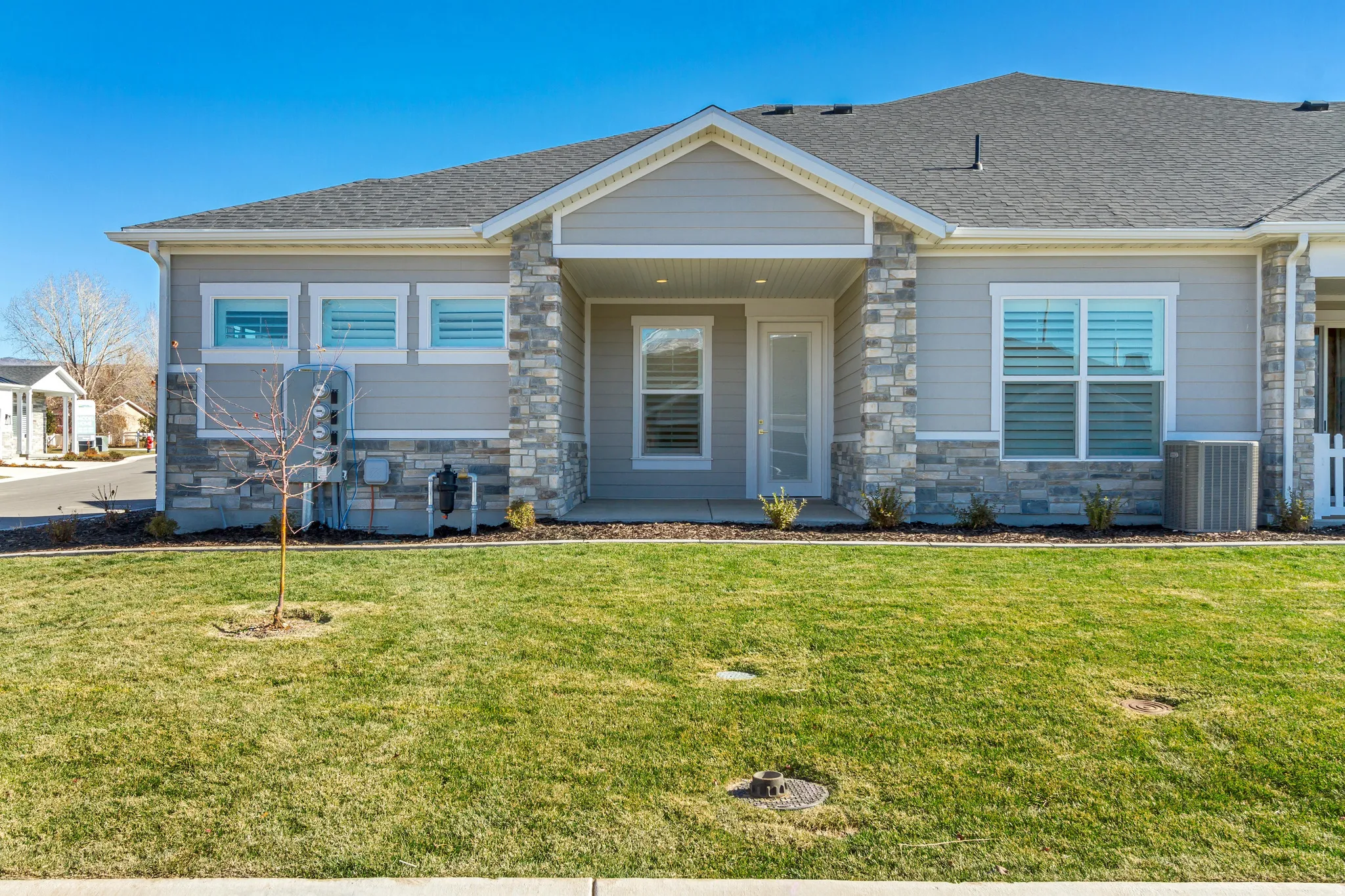 Contemporary exterior featuring light gray siding, stone veneer accents, an inviting covered entry, a prominent front window with louvered blinds, and a well-maintained lawn