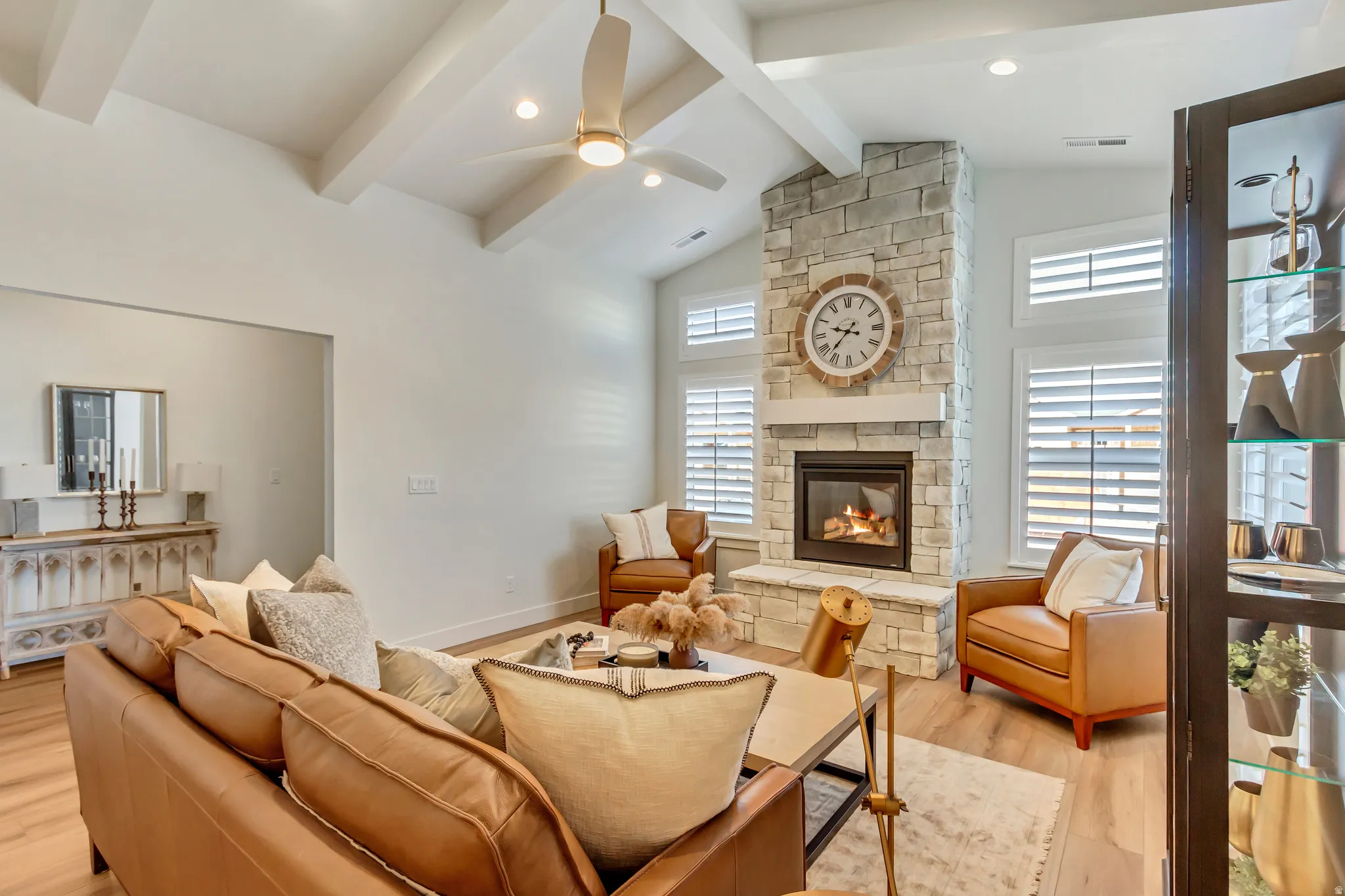 Living area featuring a stone-clad fireplace with a white mantel, vaulted ceilings with exposed beams, wood-finish flooring, and white plantation shutters on multiple windows