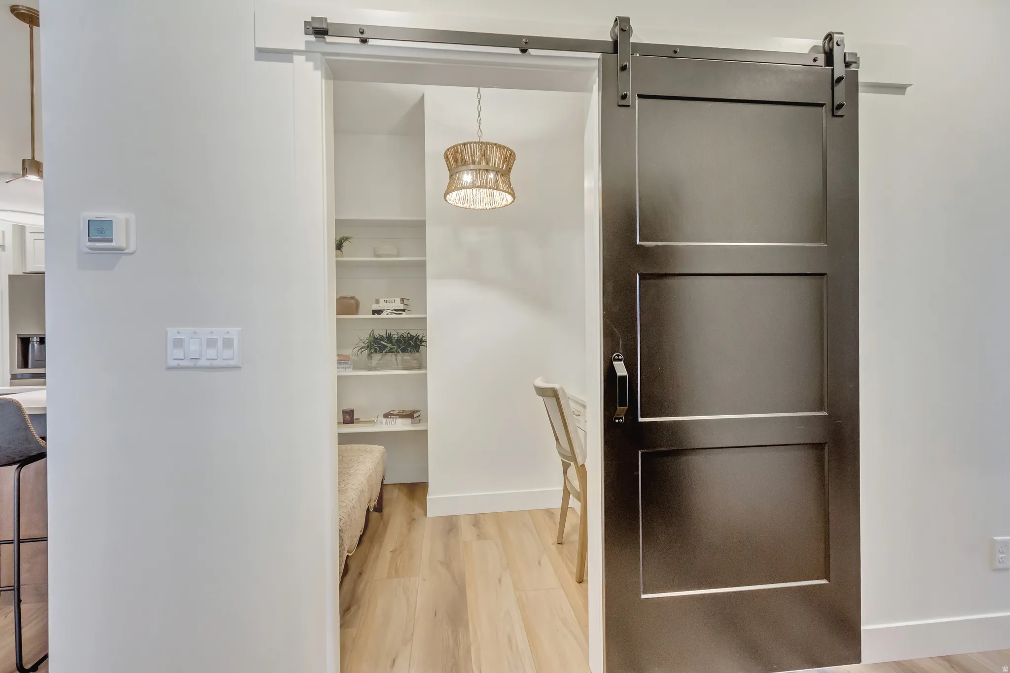 Dark-toned barn door with metal hardware providing access to an interior space featuring built-in shelving, wood-finish flooring, and a woven pendant light fixture