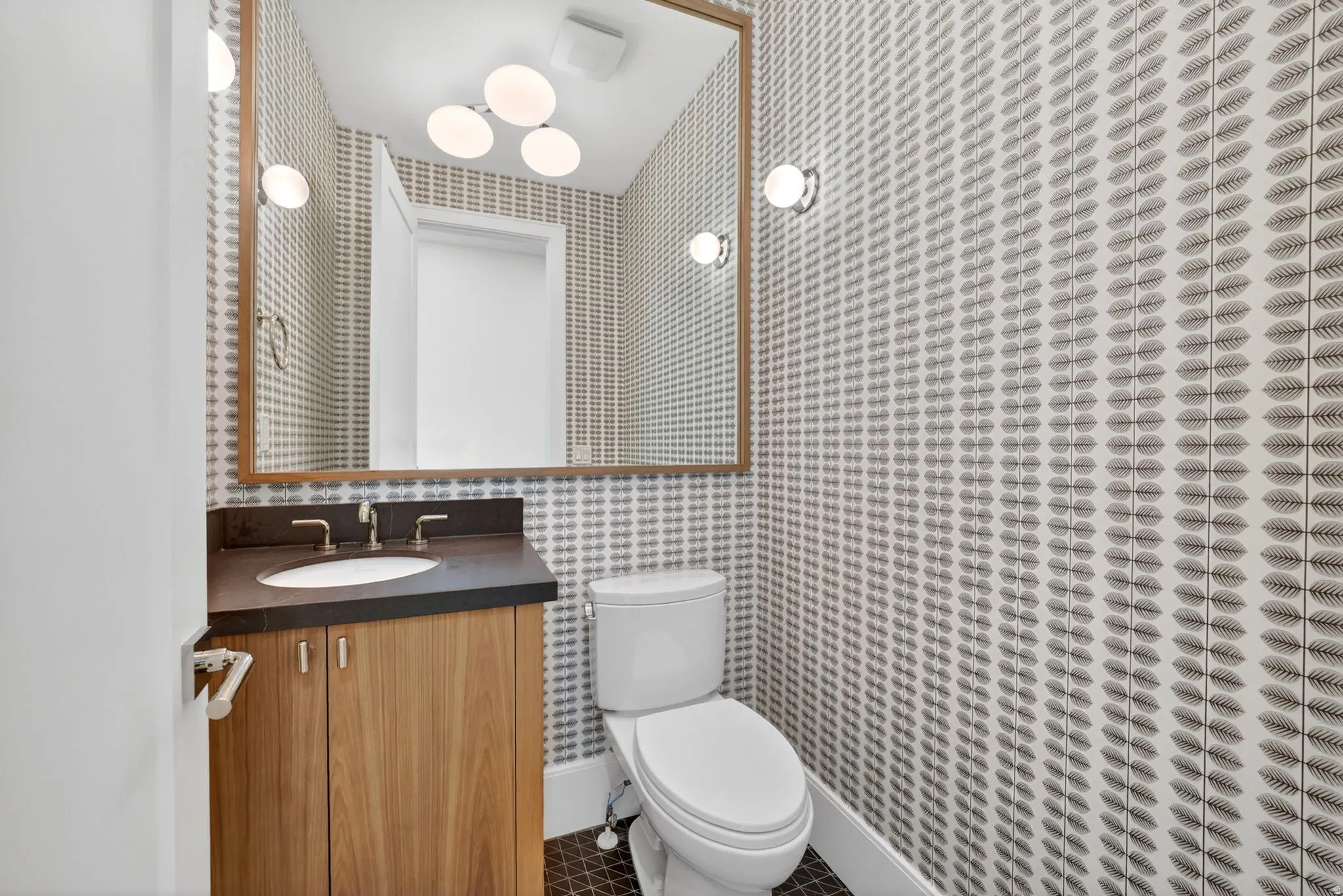 Powder room featuring a single-sink vanity with wood-finish cabinetry and a dark countertop, framed mirror, and patterned wallpaper