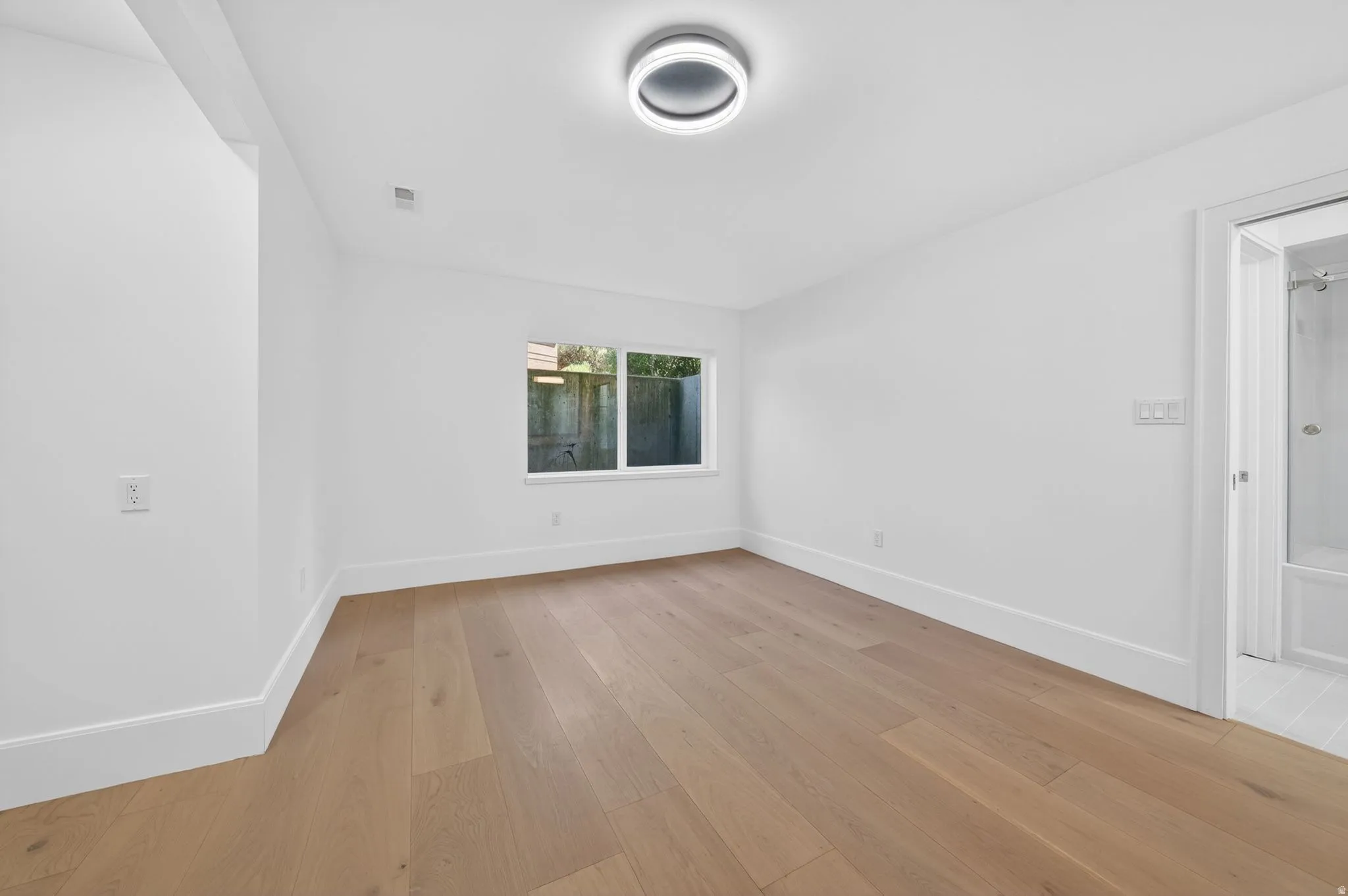 Bright room featuring wood-finish flooring, white walls, a window, a contemporary flush-mount ceiling light, and an entry to a bathroom with a shower-over-tub combination