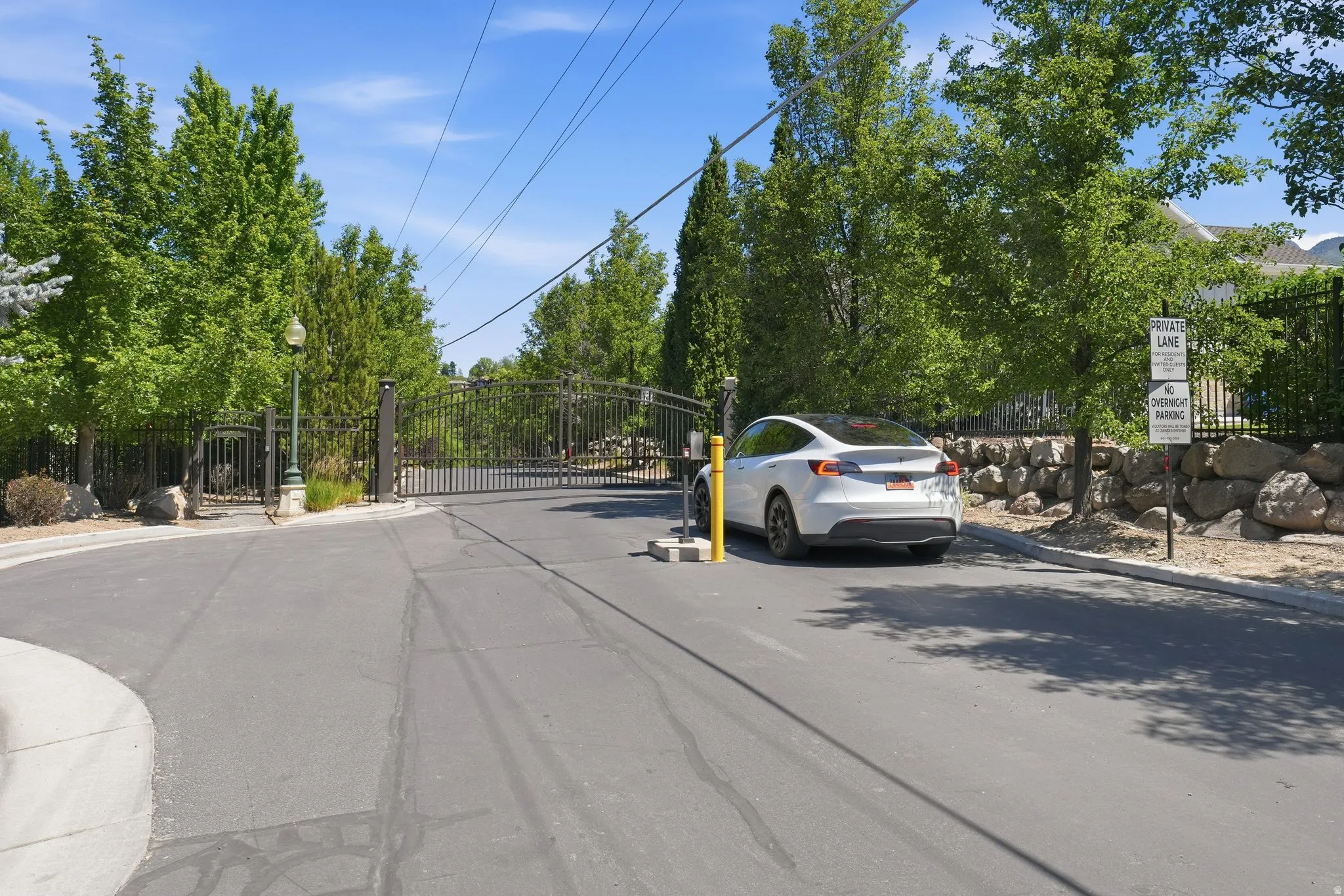 Gated entry featuring ornate wrought iron gates, mature landscaping, a stone retaining wall, and a paved private lane