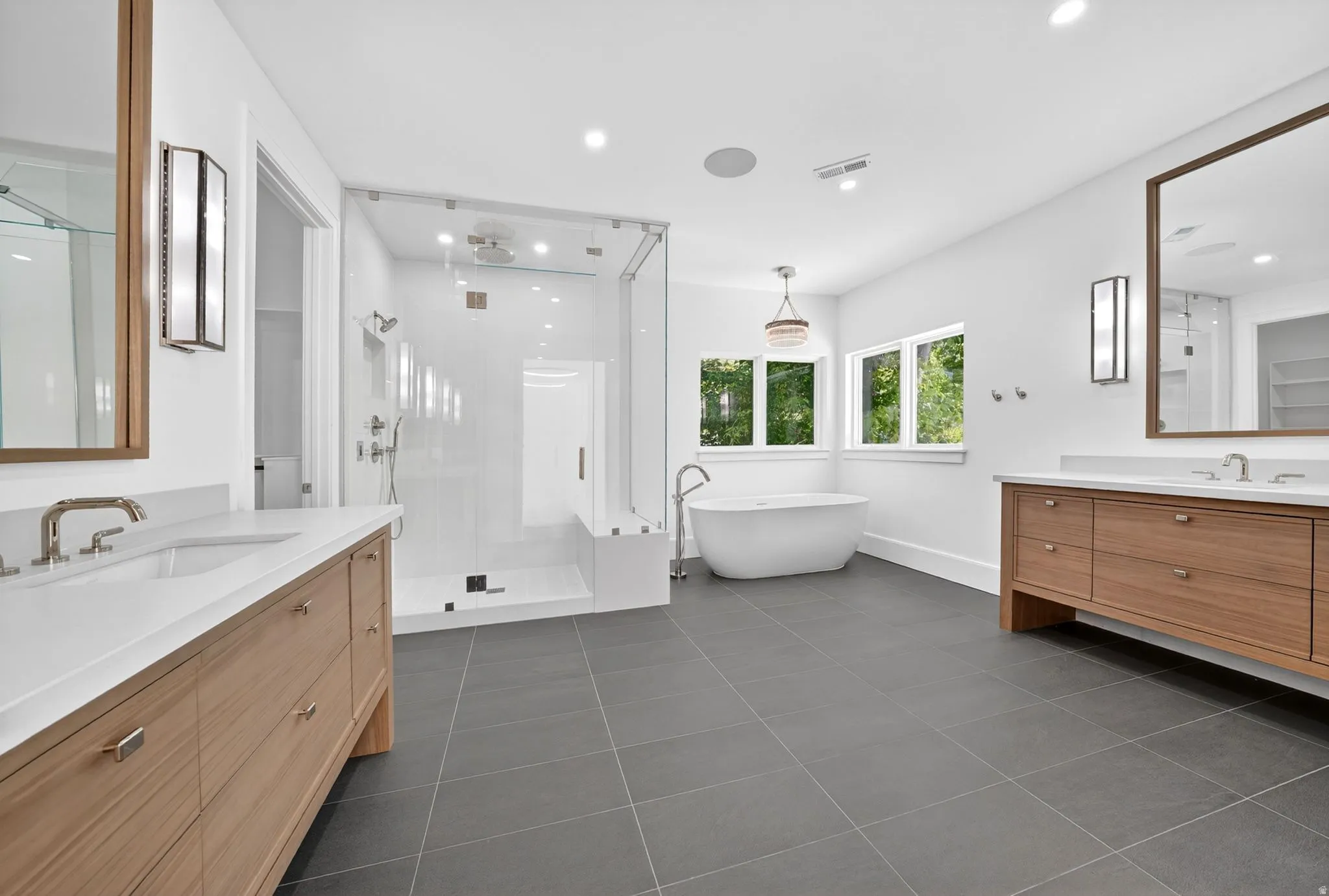 Modern bathroom featuring dual wood-finish vanities with white countertops, a freestanding soaking tub, a frameless glass shower enclosure with a built-in bench, and large format tile flooring