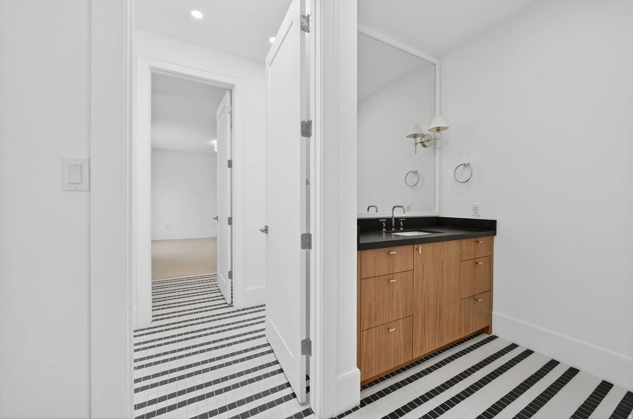 Contemporary vanity area featuring a black and white striped tile floor, a light wood-finish vanity with a dark countertop, a rectangular mirror, and sconce lighting