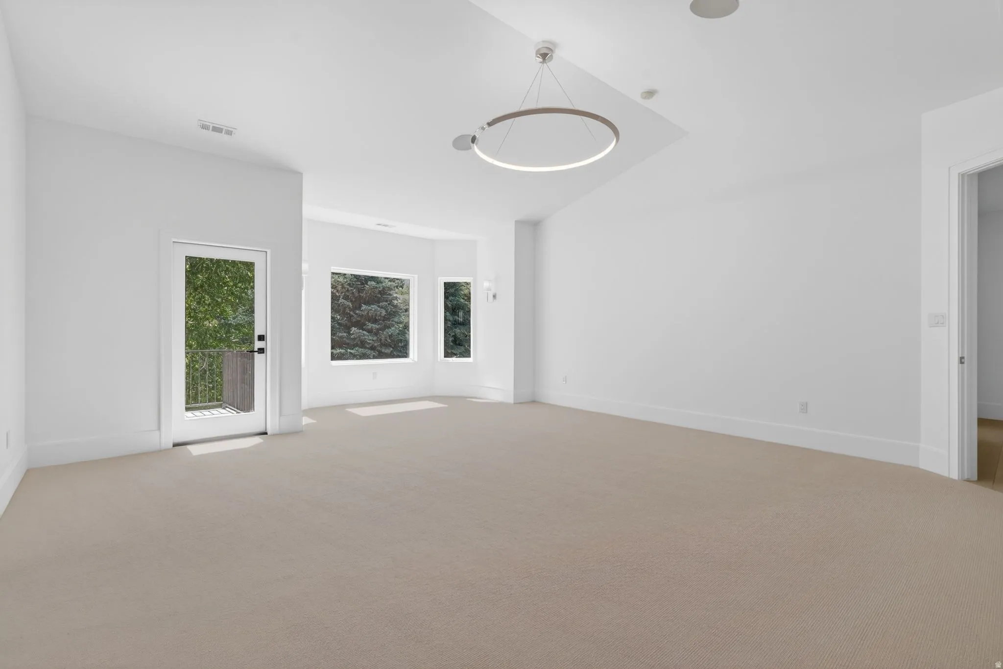 Spacious primary bedroom featuring neutral carpeting, a modern circular ceiling light fixture, multiple windows, and a glass-paneled door opening to an exterior balcony